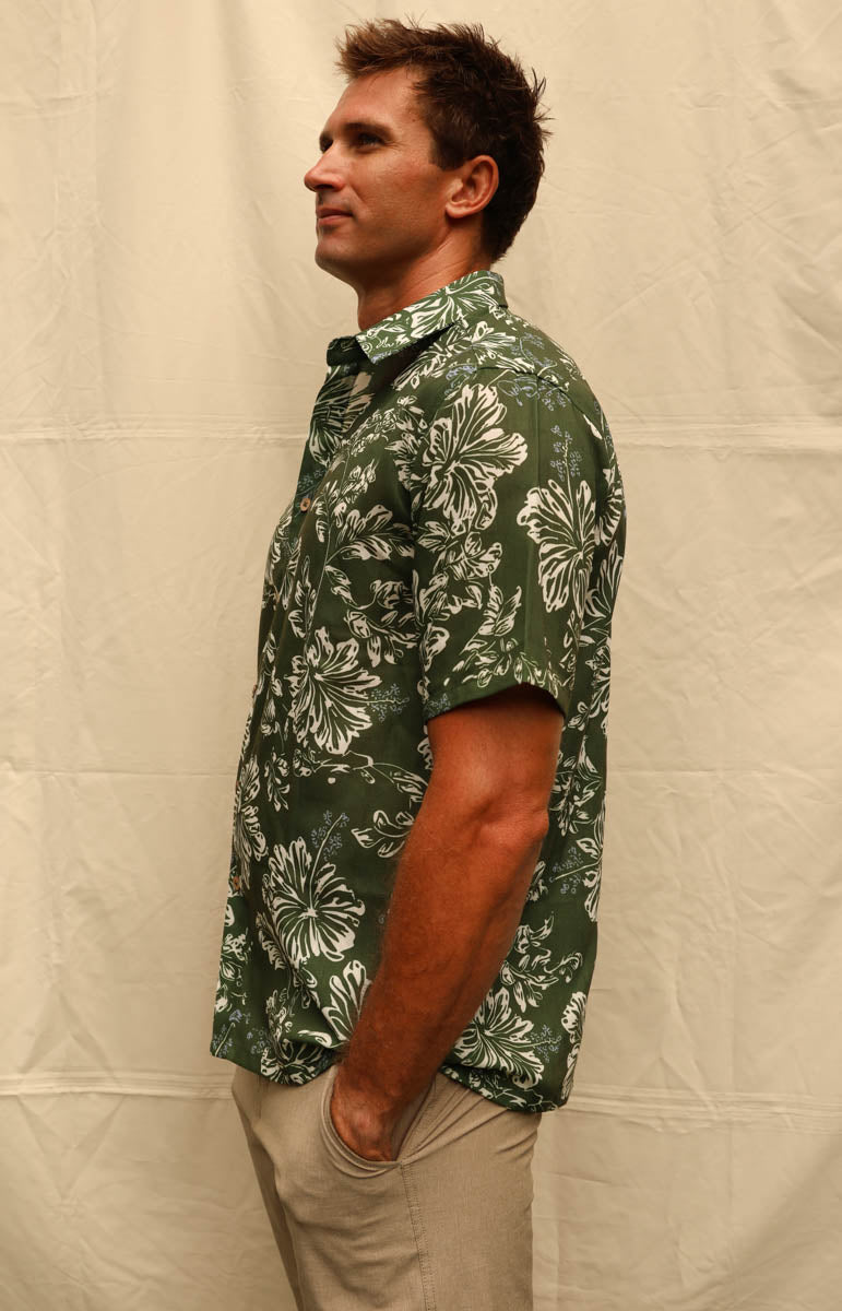 A man wearing a green short-sleeve Hawaiian button-down shirt with a white floral pattern, paired with khaki pants, standing against a beige fabric backdrop. Tai swim braden shirt in kaimana aloha shirt hemp lyocell button down