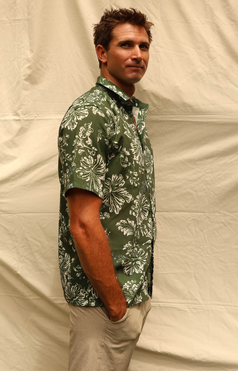 A man wearing a green short-sleeve button-up shirt with a white floral pattern, paired with khaki pants, standing against a wrinkled beige backdrop.
