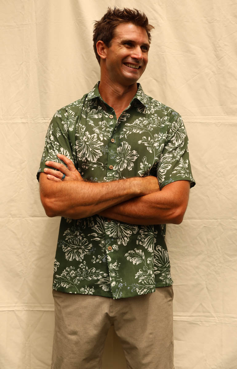 A man wearing a green short-sleeve button-up shirt with a white floral pattern and khaki pants, standing with arms crossed against a beige fabric backdrop. tai swim braden shirt in kaimana aloha shirt hemp lyocell button down