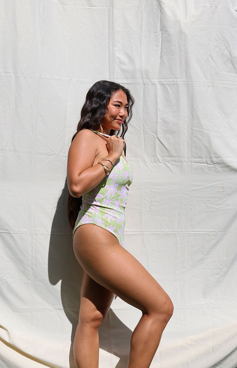 Woman in a green swimsuit standing against a white backdrop Woman wearing a green and pink floral one-piece swimsuit against a white backdrop tai swim co ella one piece in hawi green and pink light floral trippy pattern from oahu kailua sustainable bikinis hawaiian designed swimsuits
