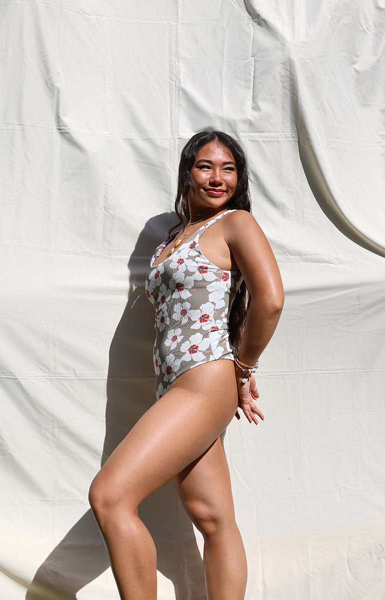 tai swim co ella one piece in kailua brown one piece with white floral hibiscus flowers scattered on ribbed sustainable fabric scoop neck bikini top and bottom from kailua oahu hawai'i on a sunny day bright model sunshine outdoor on white backdrop from hawaii