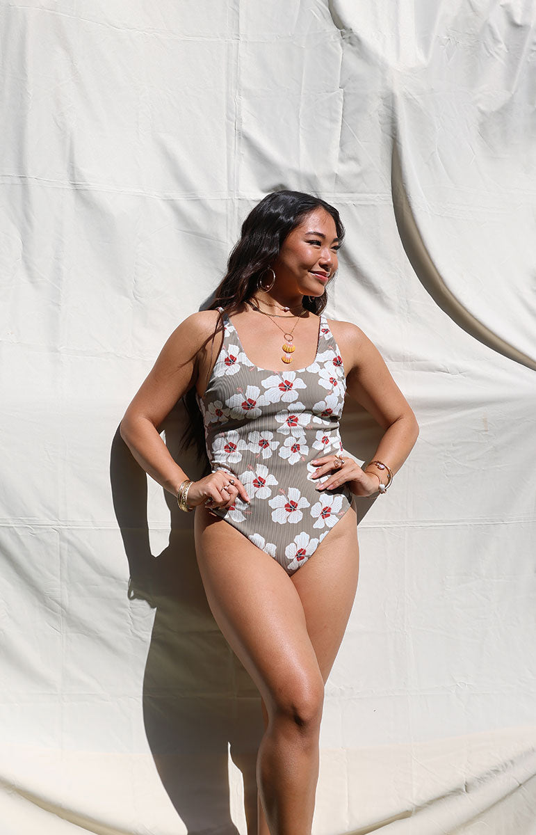 tai swim co ella one piece in kailua brown one piece with white floral hibiscus flowers scattered on ribbed sustainable fabric scoop neck bikini top and bottom from kailua oahu hawai'i on a sunny day bright model sunshine outdoor on white backdrop