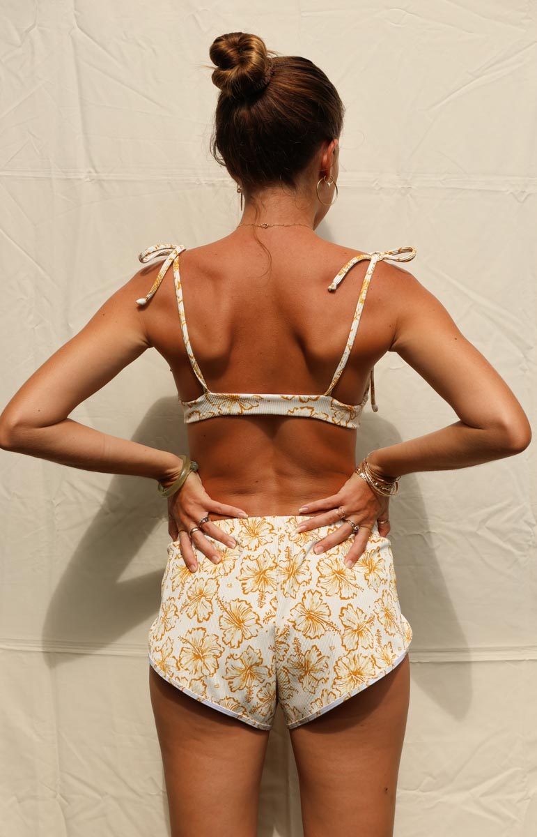tai swim waimea yellow hibiscus print swimwear from oahu kailua hawaii Woman wearing a floral two-piece outfit against a textured beige wall.