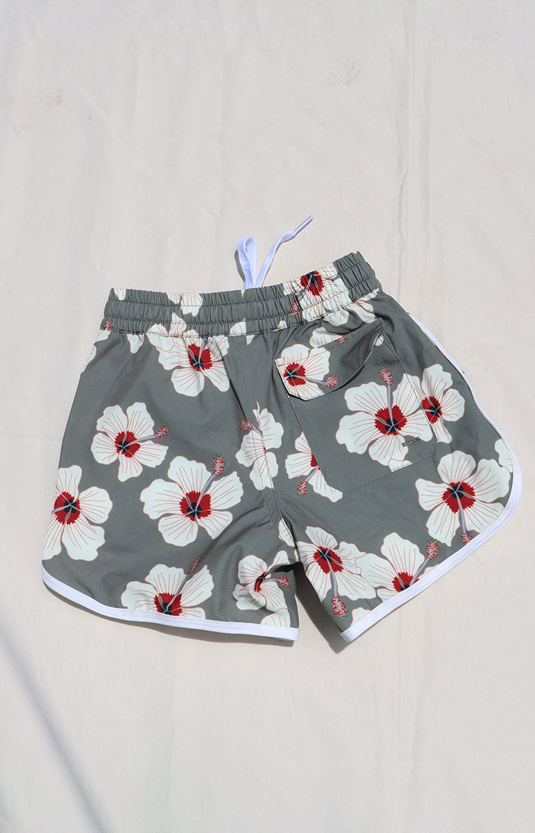 Tai Swim Co Keiki Keahi boardshorts swim trunks in Kailua hibiscus print, made with eco-conscious fabric and UPF protection. Features include a soft elastic waistband with white contrast drawstring, curved side seams with white trim, and a functional back patch pocket. Designed for comfort and sun safety, available in kids sizes 1/2 through 10/12. Pictured laid flat on a sunlit neutral background in hawaii with back pocket