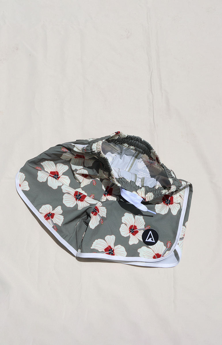 Tai Swim Co Keiki Keahi boardshorts swim trunks in Kailua hibiscus print, made with eco-conscious fabric and UPF protection. Features include a soft elastic waistband with white contrast drawstring, curved side seams with white trim, and a functional back patch pocket. Designed for comfort and sun safety, available in kids sizes 1/2 through 10/12. Pictured laid flat on a sunlit neutral background in hawaii