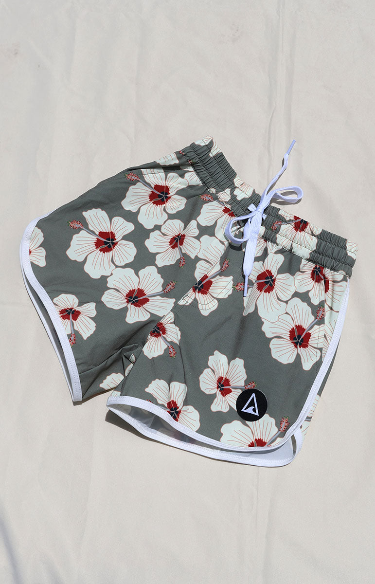 Tai Swim Co Keiki Keahi boardshorts swim trunks in Kailua hibiscus print, made with eco-conscious fabric and UPF protection. Features include a soft elastic waistband with white contrast drawstring, curved side seams with white trim, and a functional back patch pocket. Designed for comfort and sun safety, available in kids sizes 1/2 through 10/12. Pictured laid flat on a sunlit neutral background in hawaii