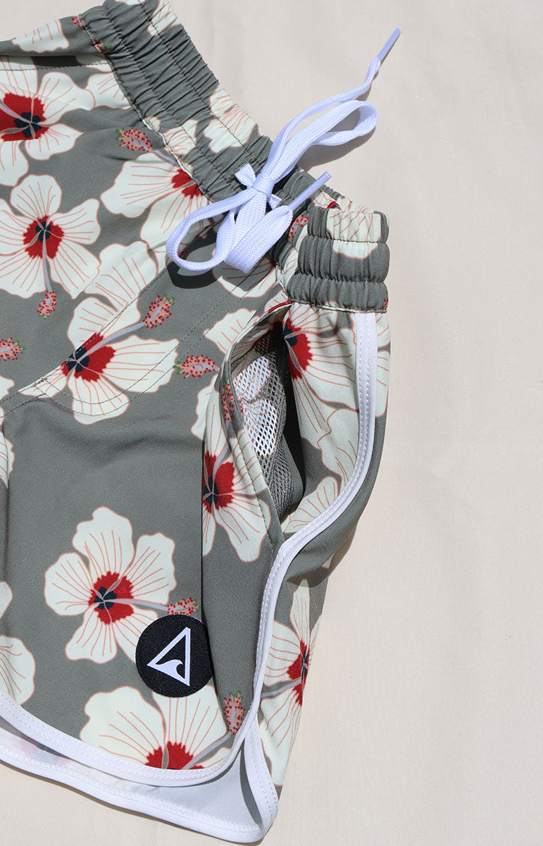 Tai Swim Co Keiki Keahi boardshorts swim trunks in Kailua hibiscus print, made with eco-conscious fabric and UPF protection. Features include a soft elastic waistband with white contrast drawstring, curved side seams with white trim, and a functional back patch pocket. Designed for comfort and sun safety, available in kids sizes 1/2 through 10/12. Pictured laid flat on a sunlit neutral background in hawaii wish mesh inside