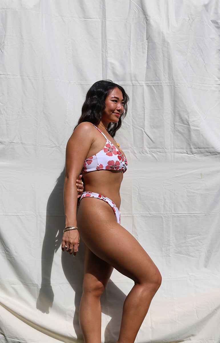 tai swim co cheeky thong bikini bottom in haleiwa red floral hibiscus color print from oahu kailua hawaii minimal coverage bikini set from sustainable hawaiian designer tai swim in kailua on model with dark hair and bralette skimpy top and sunrise shell necklace