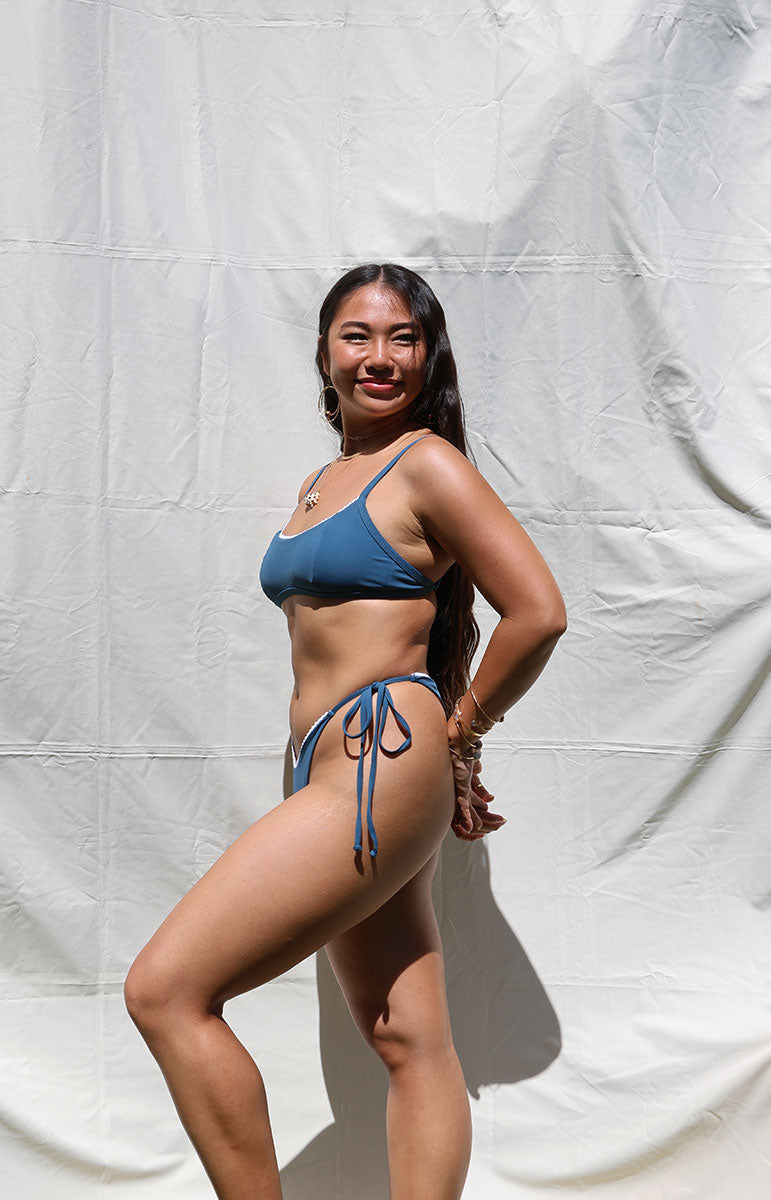 tai swim co rhia top in rift dark Blue sporty scoopneck with a lace detail, side tie sliding adjustable, sliding straps, scoop neck back Hawaiian swimwear from Oahu Kailua, Hawaii with matching adjustable side tie, stringy high waisted bottom with leasing pipe border inspired by dark blue ocean waves navy blue, dark blue bikini with contrast, border, piping and shell model necklace with dark hair
