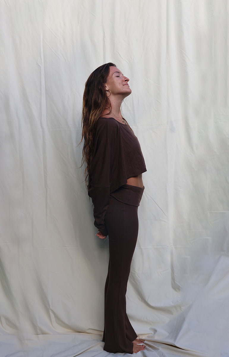 Woman in brown outfit standing against a white curtain Woman wearing a brown outfit standing against a white backdrop tai swim co shaiann top long sleeve crop top in espresso