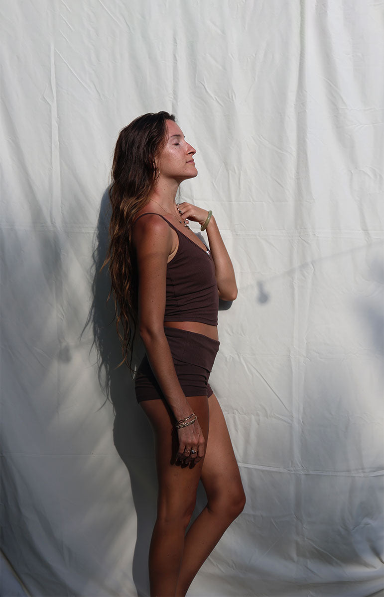 Woman in a brown outfit standing against a white backdrop Woman wearing a brown tank top and shorts against a white background tai swim co shannah tank top in espresso brown adjustable top and foldover shorts from oahu kailua hawaii