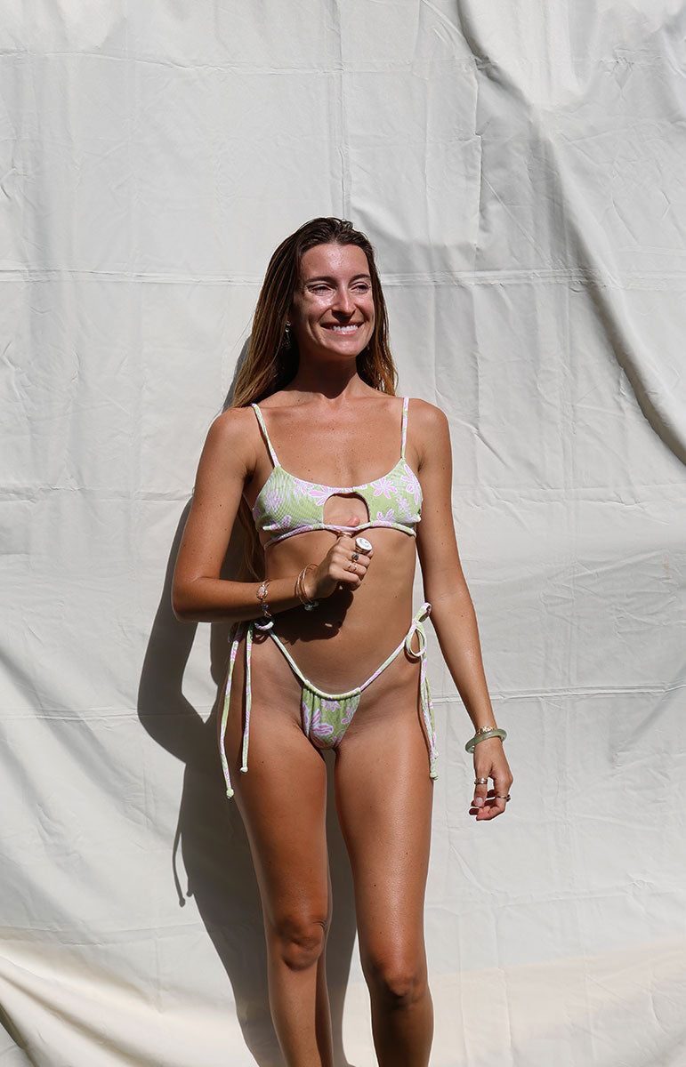 Woman in a bikini standing against a white curtain cheeky side tie tai bottom in hawi grene and pink floral stringy thong bikini bottom brazilian style cheeky cut swimsuit bottoms from hawaii tai swim co