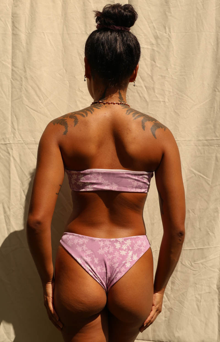 Rear view of a woman wearing a light purple bikini with a floral pattern, standing against a beige fabric backdrop. Tai Swim Manoa Purple Donna Top Donna Bottom Bikinis