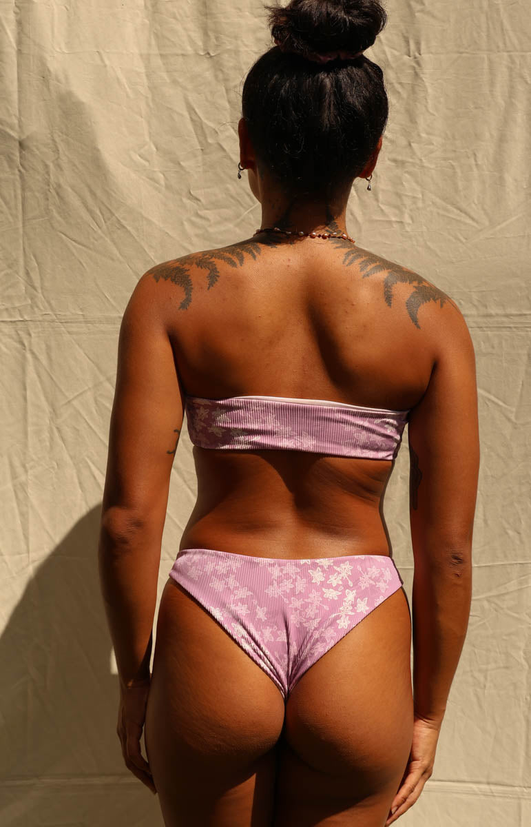 A woman viewed from behind wearing a light pink bikini with a white floral pattern, standing against a wrinkled beige backdrop.
