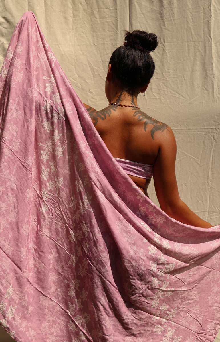 A woman shown from behind wearing a pink satin-like bandeau top and holding a large pink fabric drape with a subtle white floral pattern, set against a wrinkled beige backdrop.