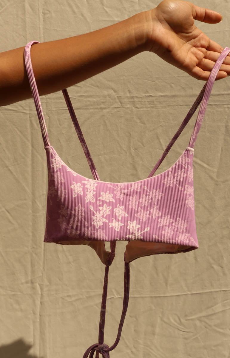 A pink ribbed knit bikini top with white floral patterns and thin shoulder straps, held up by a person's arm against a beige fabric backdrop.
