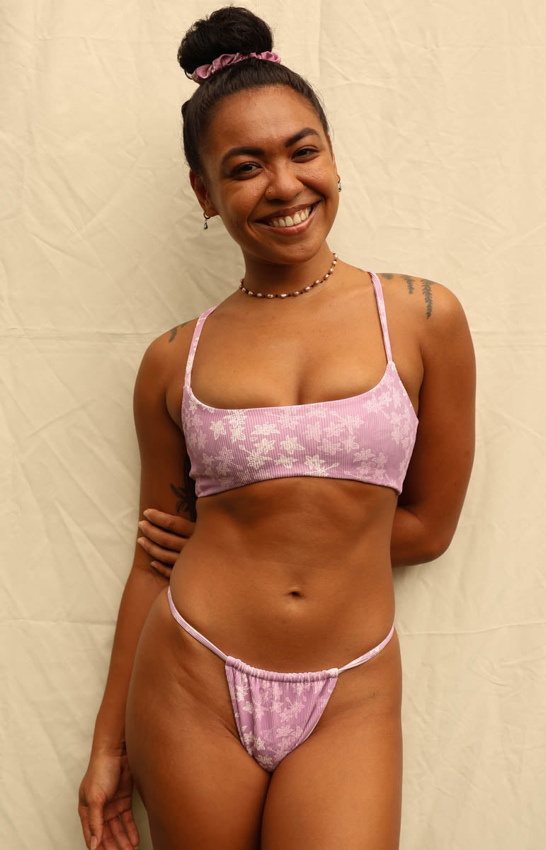 A smiling woman with her hair in a bun wears a light pink floral bikini set against a beige fabric backdrop.