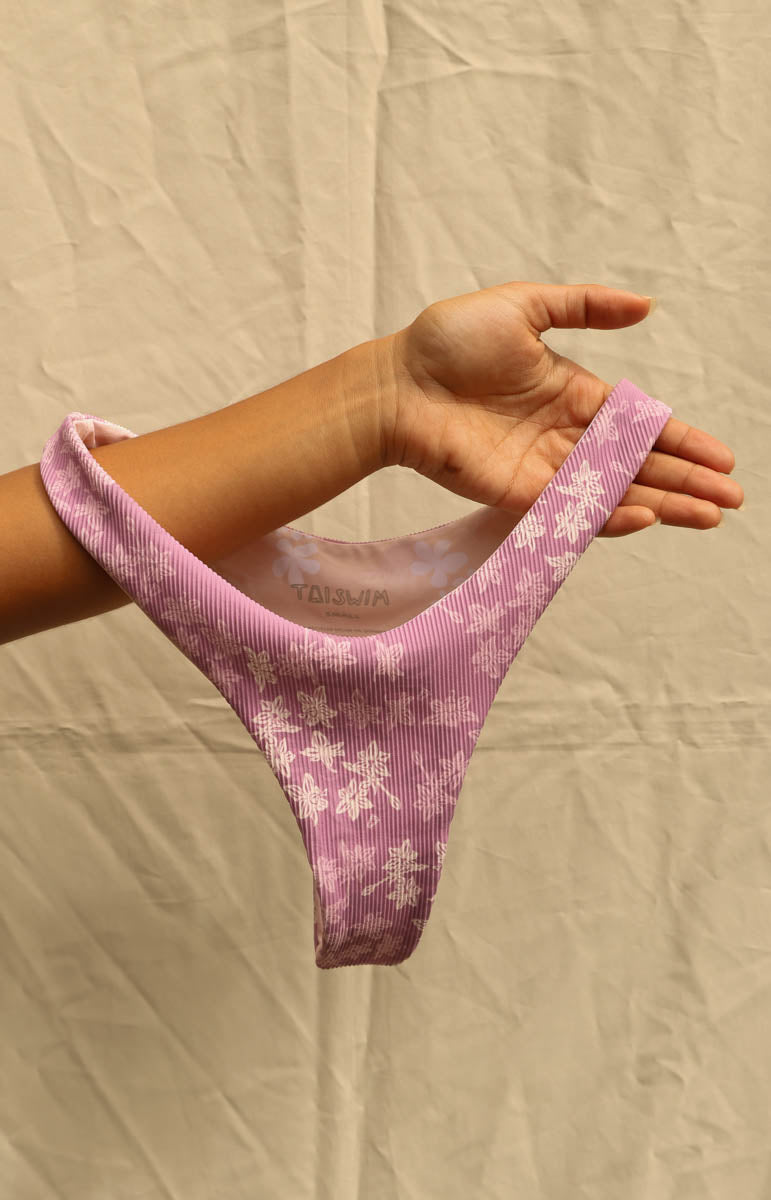 A person's hand holds up a ribbed pink bikini bottom with a white floral pattern, set against a wrinkled beige fabric background.