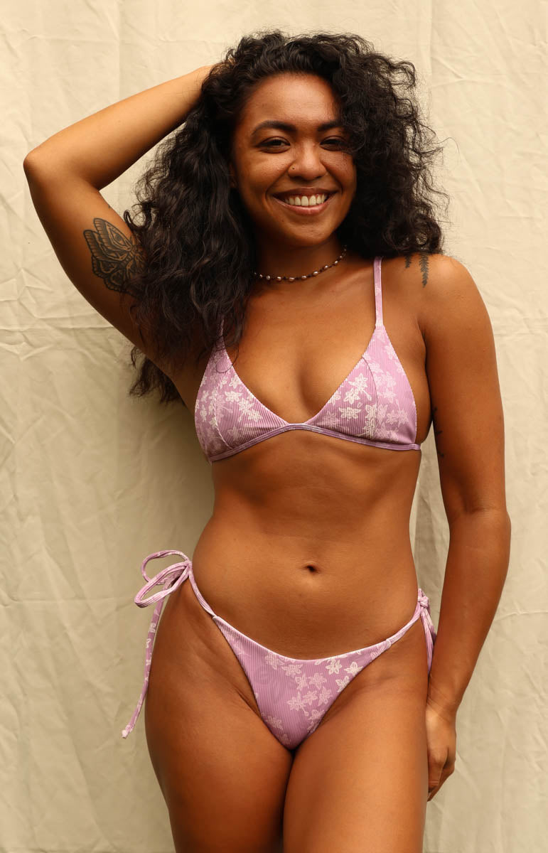 A woman modeling a light purple and white floral print string bikini against a neutral beige backdrop. Tai Swim Manoa print bikini lavender purple swimwear