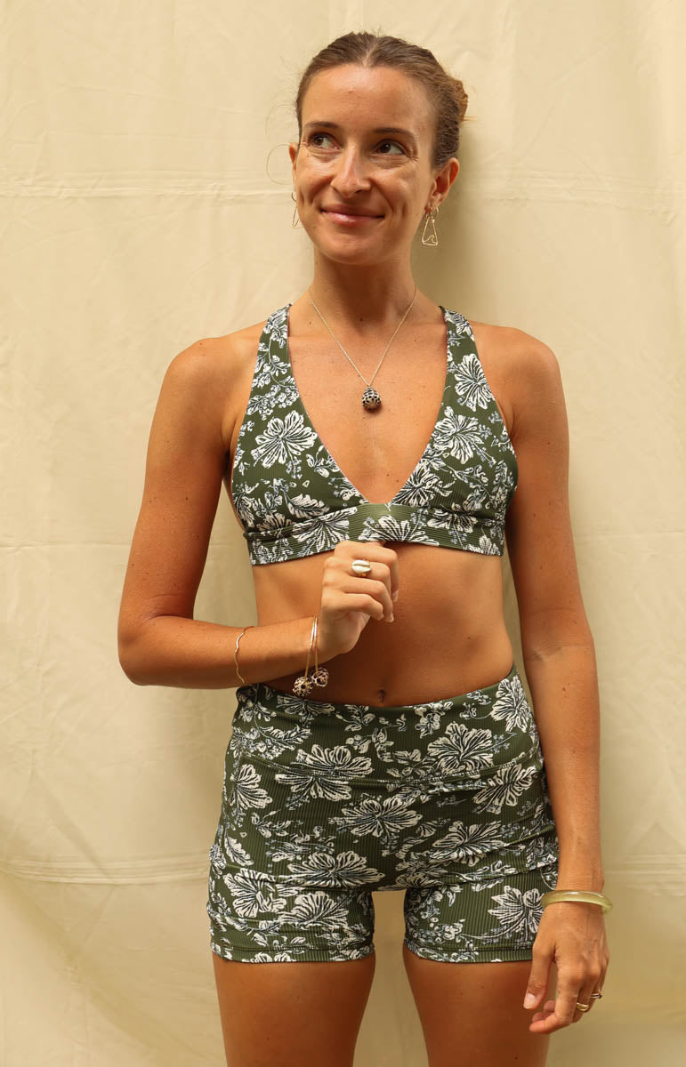 A woman shown from the back wearing a matching two-piece set with a green and white floral pattern, consisting of a halter-style sports bra and high-waisted shorts, posed against a beige fabric backdrop. Tai Swim Linda Shorts in Kaimana