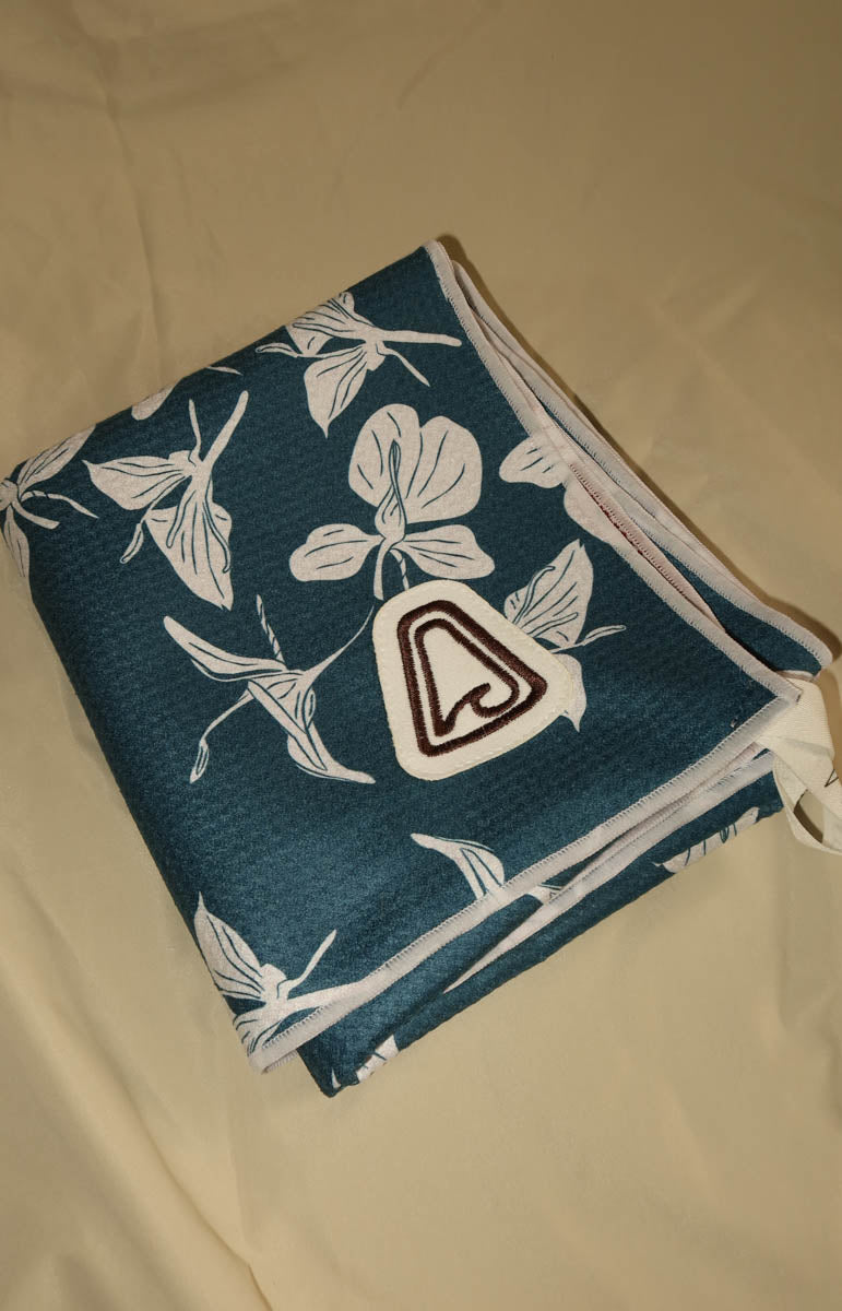 A folded dark teal fabric item with a white tropical floral print and a white patch featuring a brown stylized letter 'A', placed on a beige fabric surface. Tai Swim yoga towels sustainable