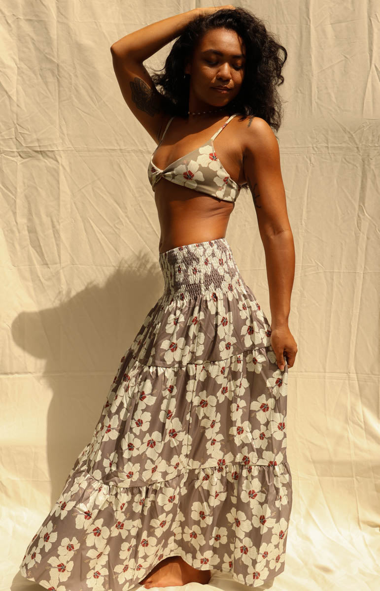 A woman modeling a matching two-piece floral set consisting of a wrap-style triangle crop top and a long tiered maxi skirt, in a gray, white, and red pattern, standing against a wrinkled beige fabric backdrop. Hibiscus floral from Kailua hawaiian brand tai swim