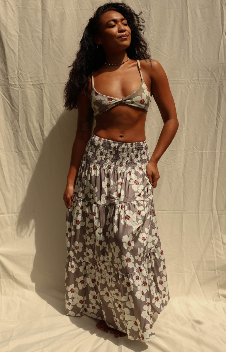 A woman modeling a matching two-piece floral set consisting of a wrap-style triangle crop top and a long tiered maxi skirt, in a gray, white, and red pattern, standing against a wrinkled beige fabric backdrop. Hibiscus floral from Kailua hawaiian brand tai swim