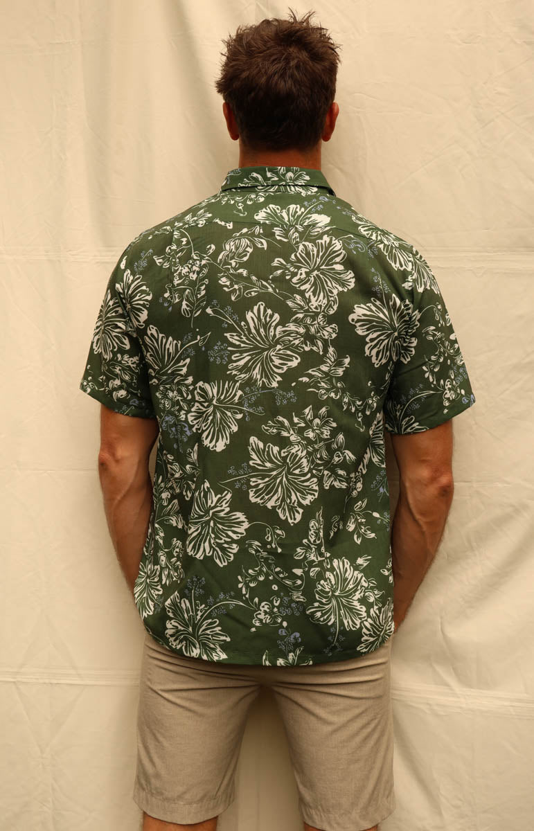 A man wearing a green short-sleeve Hawaiian button-down shirt with a white floral pattern, paired with khaki pants, standing against a beige fabric backdrop. Tai swim braden shirt in kaimana aloha shirt hemp lyocell button down