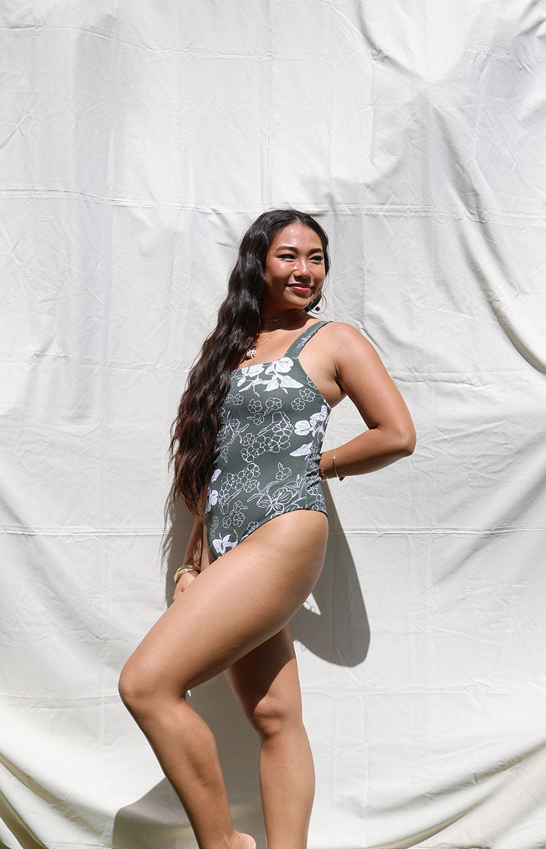 Woman wearing a tai swim green pua print green floral one-piece swimsuit against a white backdrop in hawaii cheeky reversible swimwear