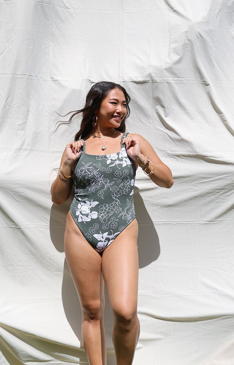 Woman wearing a tai swim green pua print green floral one-piece swimsuit against a white backdrop in hawaii cheeky reversible swimwear