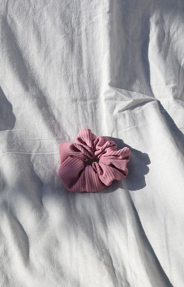 A close-up photograph of a ribbed swimwear material scrunchie in the color 'Mokihana.' The fabric has a textured ribbed pattern, featuring a blend of vibrant pink and purple hues. The colors create a gradient effect, transitioning from a deep magenta at one end to a lighter lilac towards the other, giving it a beautiful and appealing aesthetic from hawaii in kailua