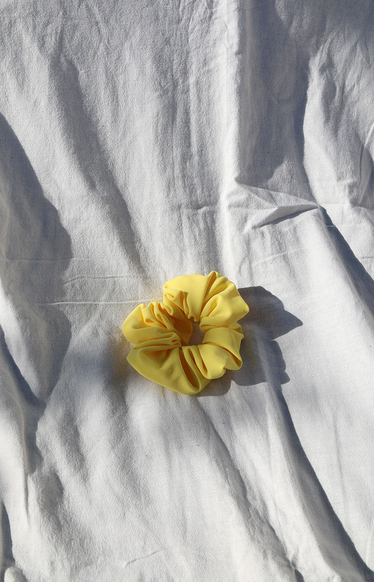 A close-up image of a ribbed swimwear scrunchie in the color Ilima yellow Oahu flower, a beautiful vibrant shade reminiscent of tropical flowers. This eco-friendly scrunchie is made in Hawaii from scrap materials, promoting sustainability and a touch of island-inspired style. The Ilima hue brings to mind the vibrant petals of the native Hawaiian ilima flower, adding a pop of lively color to your ensemble.