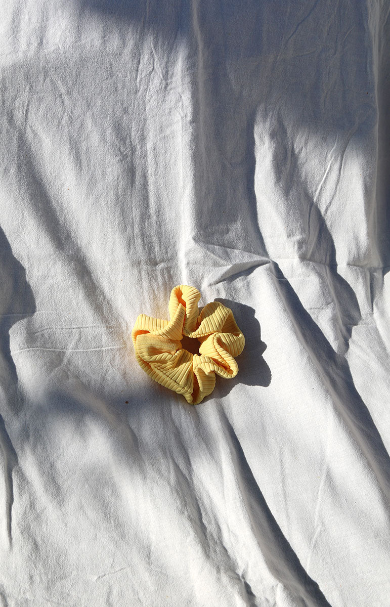 A close-up image of a ribbed swimwear scrunchie in the color Ilima yellow Oahu flower, a beautiful vibrant shade reminiscent of tropical flowers. This eco-friendly scrunchie is made in Hawaii from scrap materials, promoting sustainability and a touch of island-inspired style. The Ilima hue brings to mind the vibrant petals of the native Hawaiian ilima flower, adding a pop of lively color to your ensemble.