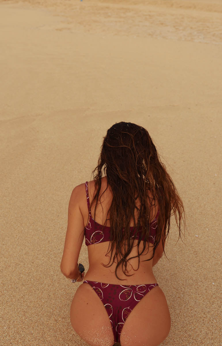 A woman with long dark hair is kneeling on a sandy beach, viewed from behind, wearing a deep maroon shell cowry tai swim bikini with a white circular pattern.