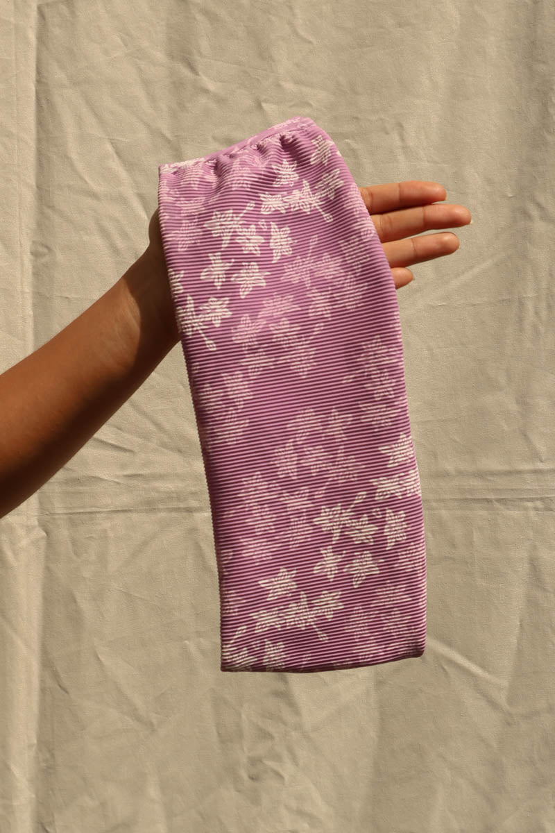 A hand holding up a lavender-purple ribbed fabric sleeve or cover with a white cherry blossom pattern, set against a wrinkled beige fabric backdrop.
