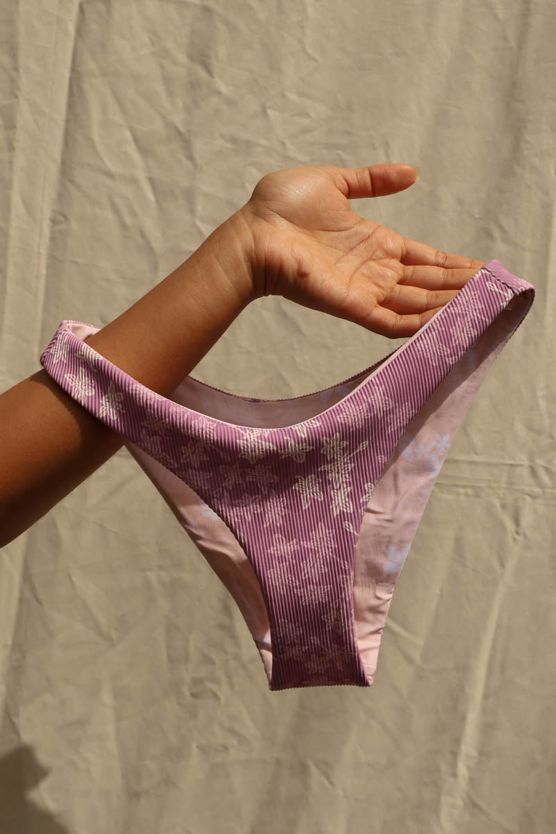 A person's hand holds up a pair of low-rise bikini bottoms with a textured, ribbed fabric in a dusty purple and light pink tie-dye pattern against a wrinkled beige backdrop. a woman wearing a light purple bikini with a floral pattern, standing against a beige fabric backdrop. Tai Swim Manoa Purple Donna Top Donna Bottom Bikinis