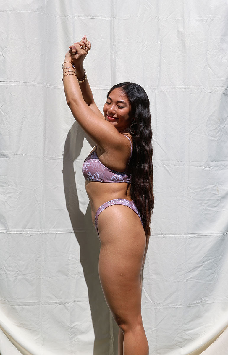 Woman wearing a purple bikini against a white curtain background in hawaii wearing tai swim co kohala smooth lining bikini drawn from hand drawn flower hibiscus from oahu kailua