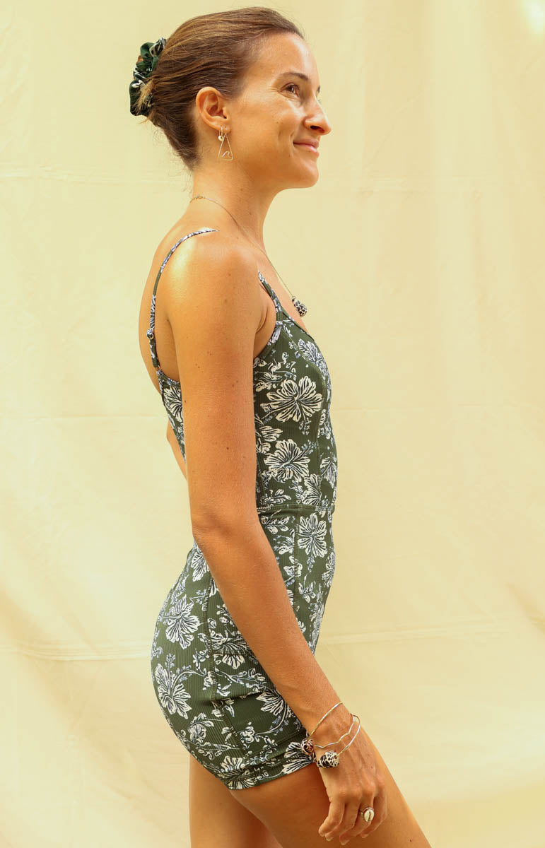 A woman modeling a green and white floral print onesie romper jumpsuit in kaimana from tai swim, standing against a light beige fabric backdrop.