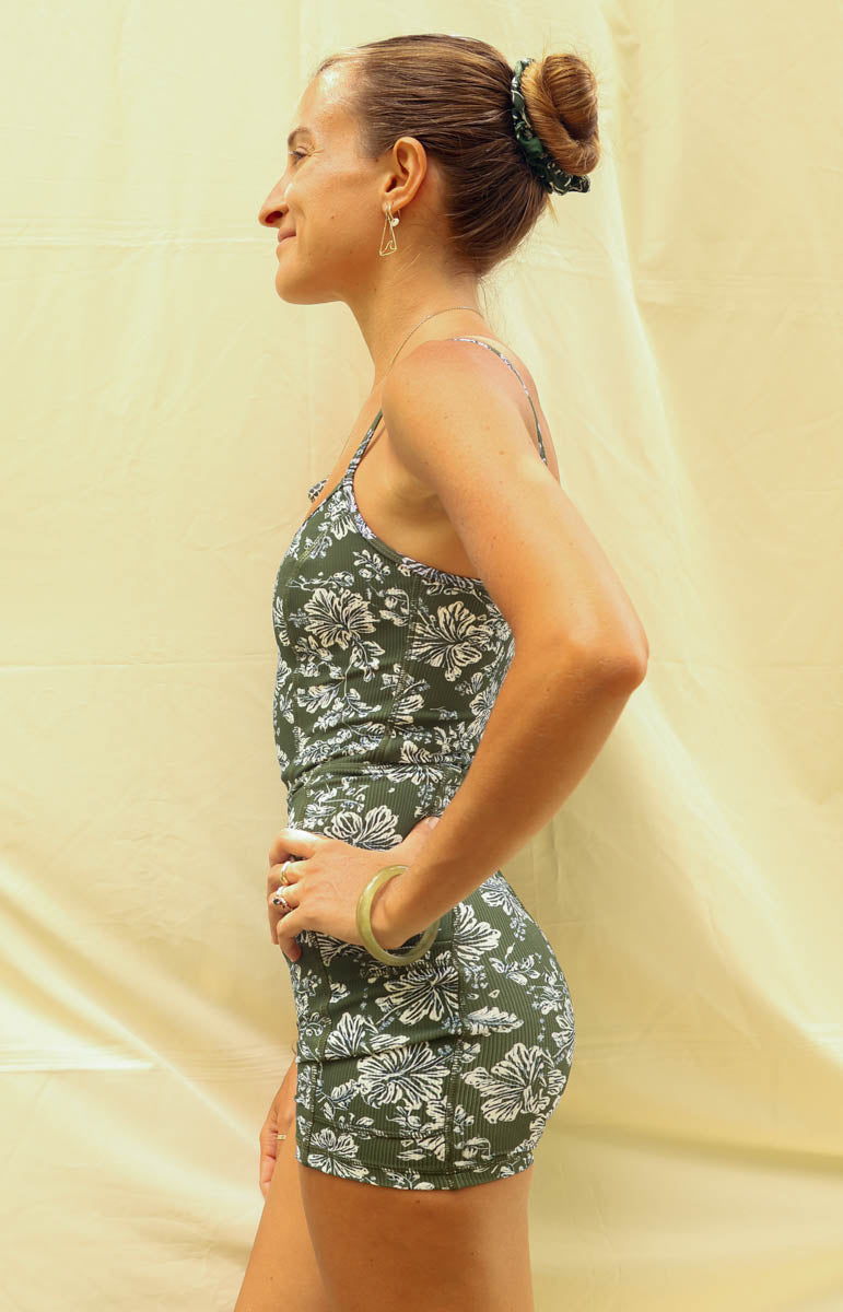 A woman modeling a green and white floral print onesie romper jumpsuit in kaimana from tai swim, standing against a light beige fabric backdrop.