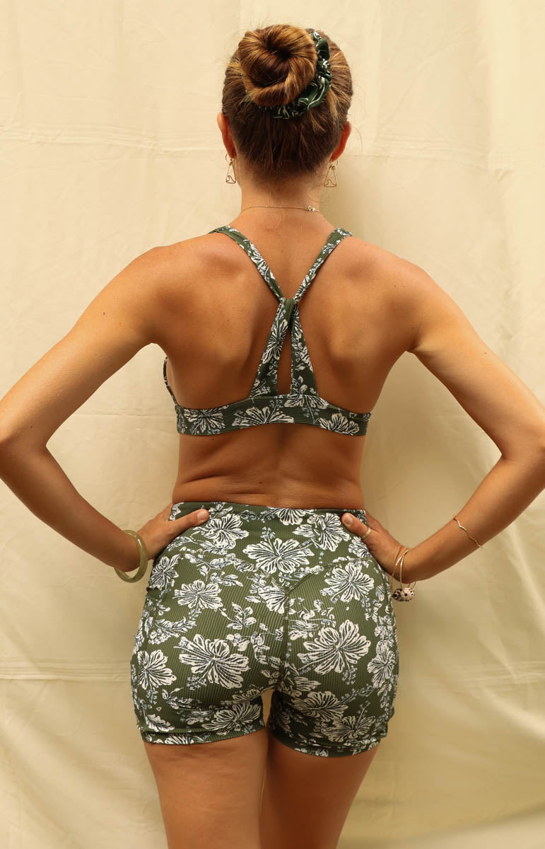 A woman shown from the back wearing a matching two-piece set with a green and white floral pattern, consisting of a halter-style sports bra and high-waisted shorts, posed against a beige fabric backdrop. Tai Swim Linda Shorts in Kaimana