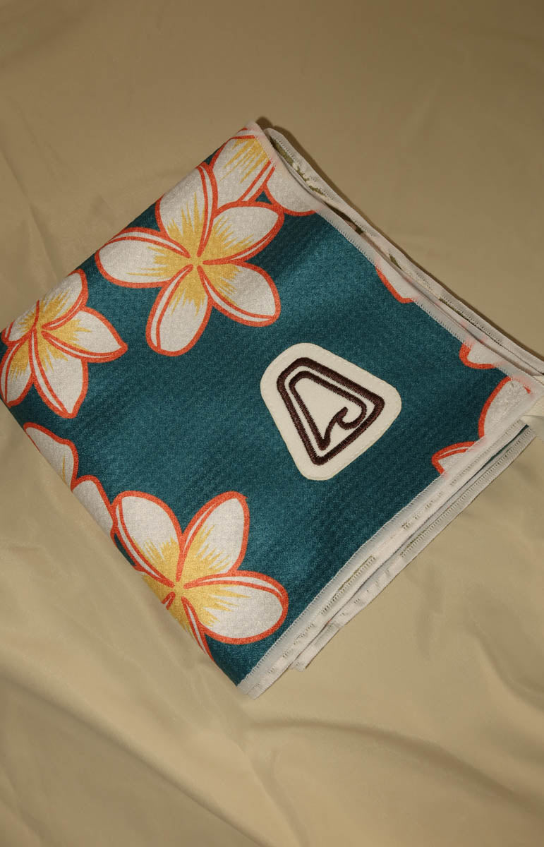 A folded teal mesh towel with a white, yellow, and orange plumeria flower pattern and a white logo patch, laid on a beige fabric background.