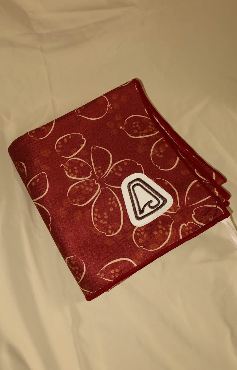 A folded red blanket with a white floral pattern and a white triangular patch featuring a black stylized logo, laid out on a white fabric background.