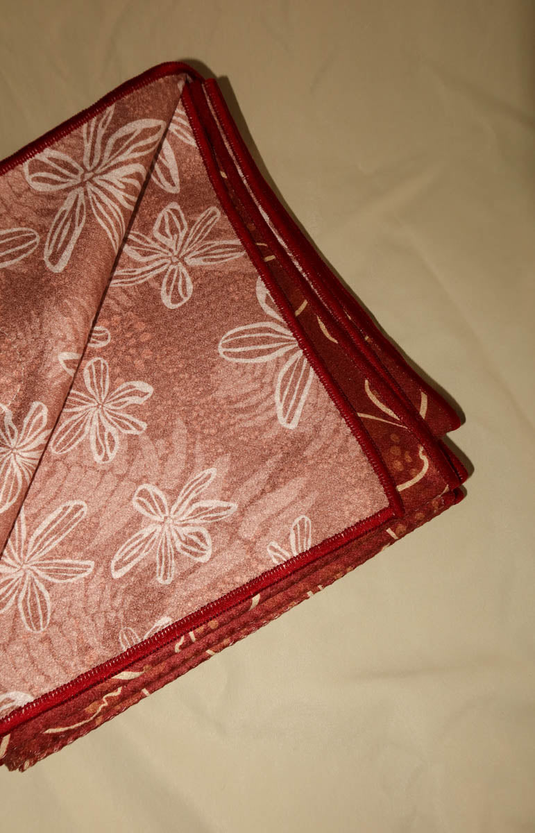 A folded red patterned fabric item, likely a pocket square or scarf, featuring a white floral print and a red satin border, laid on a beige fabric background.