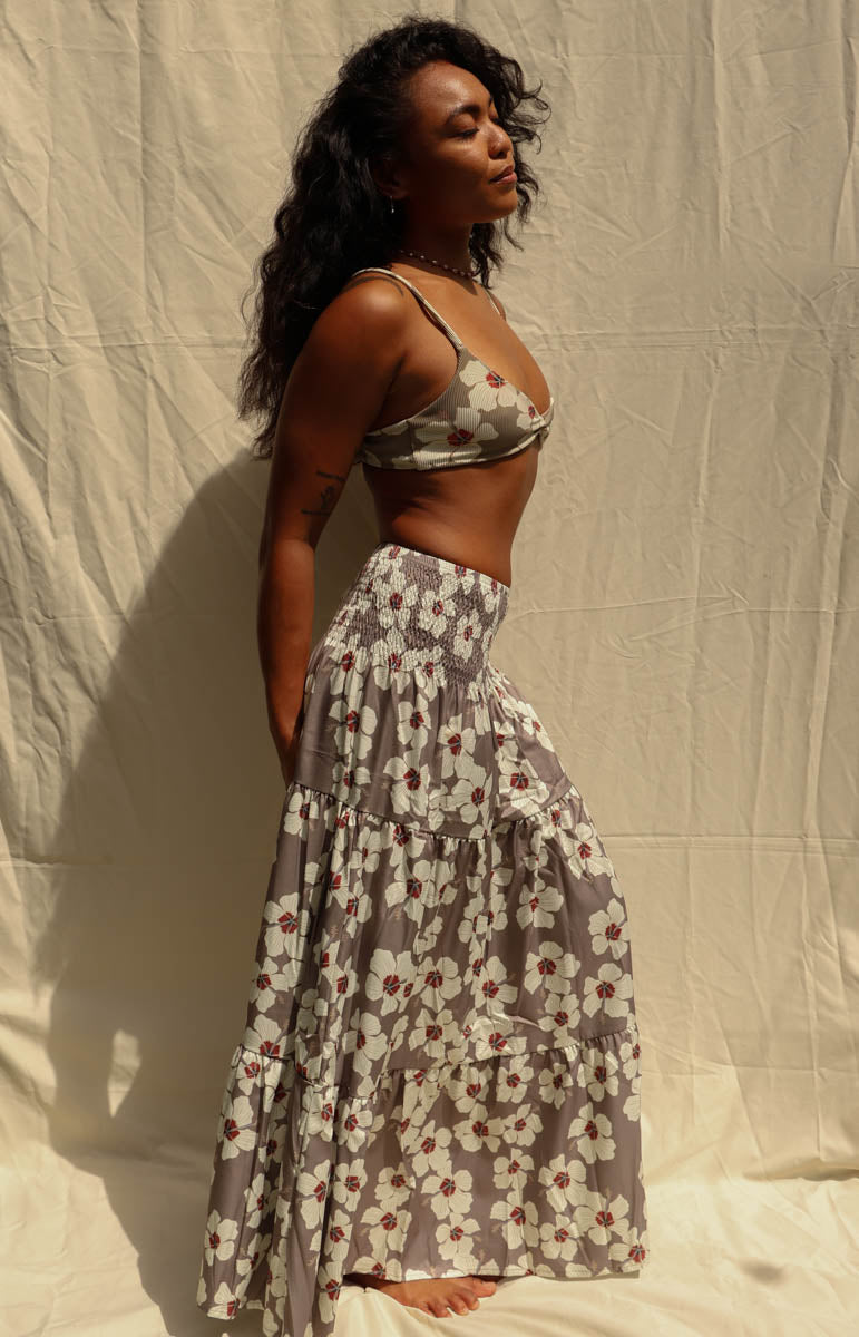 A woman modeling a matching two-piece floral set consisting of a wrap-style triangle crop top and a long tiered maxi skirt, in a gray, white, and red pattern, standing against a wrinkled beige fabric backdrop. Hibiscus floral from Kailua hawaiian brand tai swim