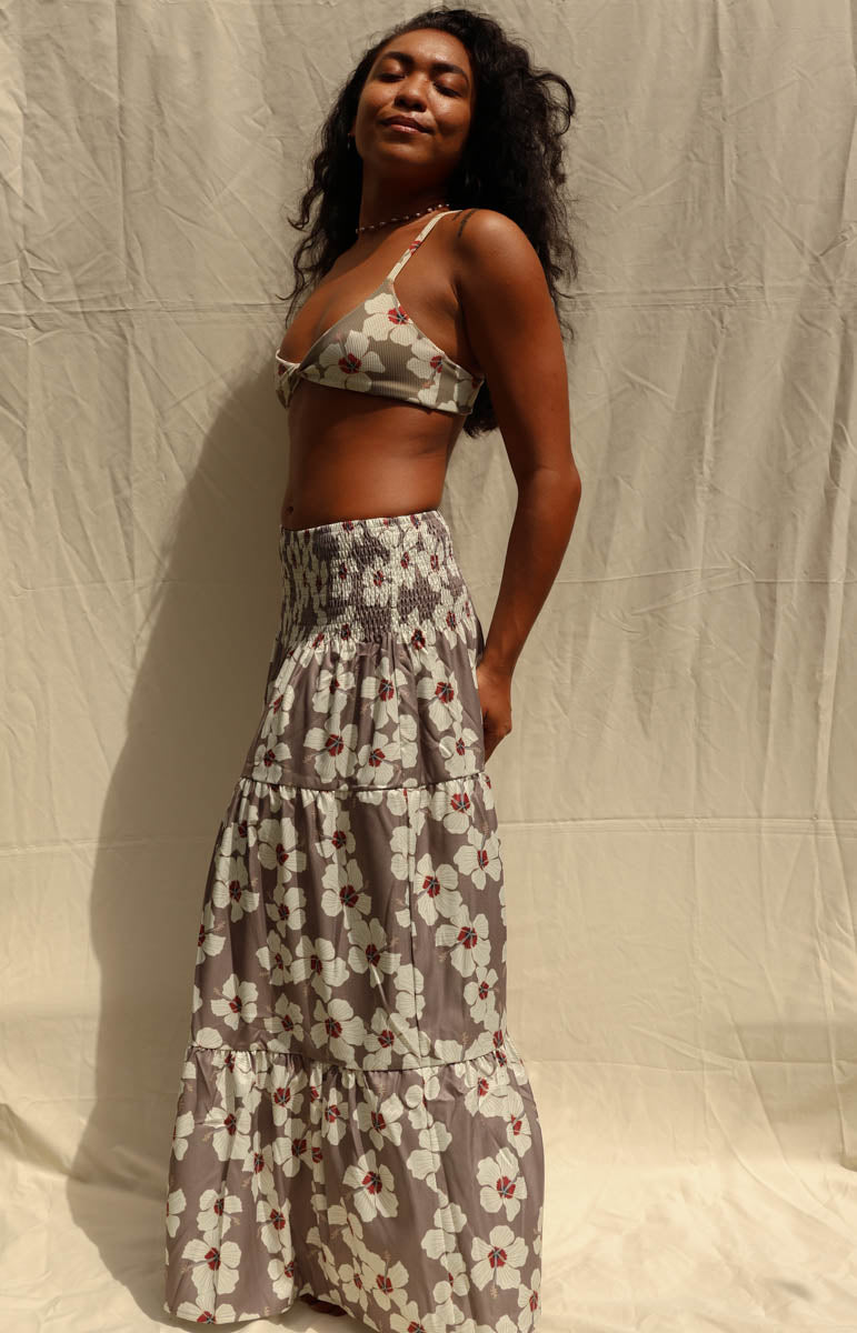 A woman modeling a matching two-piece floral set consisting of a wrap-style triangle crop top and a long tiered maxi skirt, in a gray, white, and red pattern, standing against a wrinkled beige fabric backdrop. Hibiscus floral from Kailua hawaiian brand tai swim