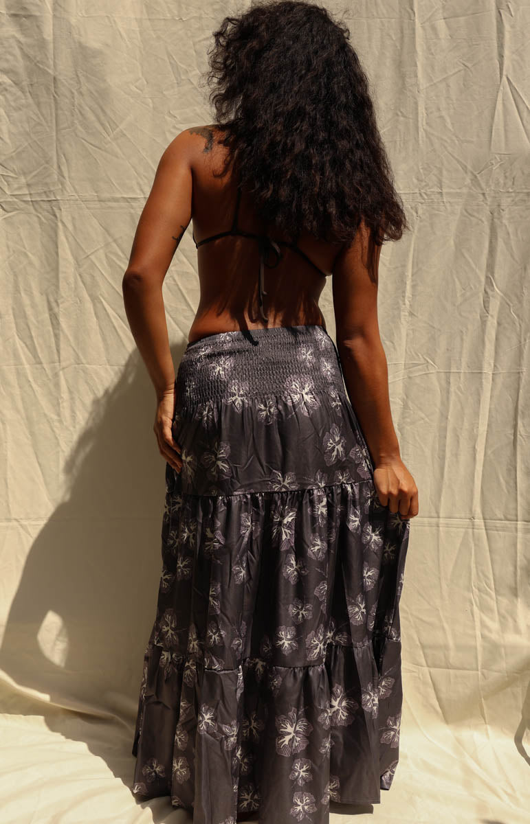 A woman modeling a dark black and gray tiered maxi skirt with a white floral pattern, paired with a black triangle bikini top, standing against a beige fabric backdrop. Tai Swim activewear skirt maxi dress