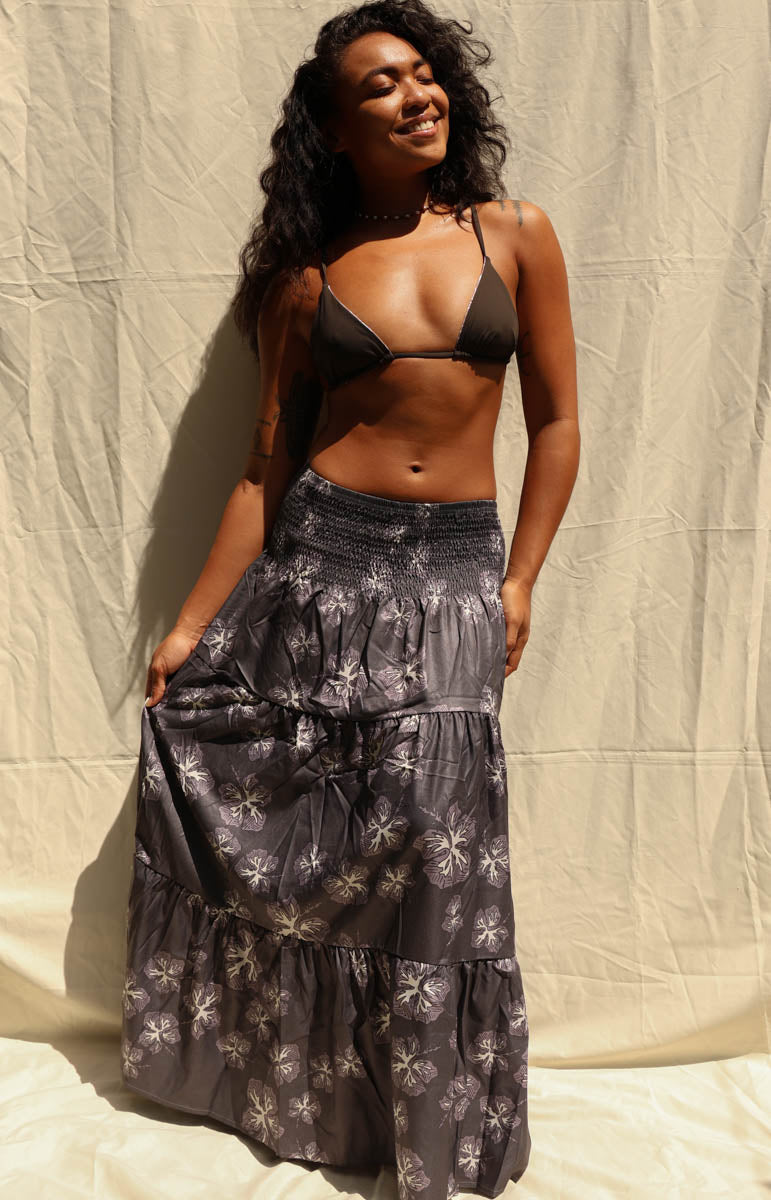A woman modeling a dark black and gray tiered maxi skirt with a white floral pattern, paired with a black triangle bikini top, standing against a beige fabric backdrop. Tai Swim activewear skirt maxi dress