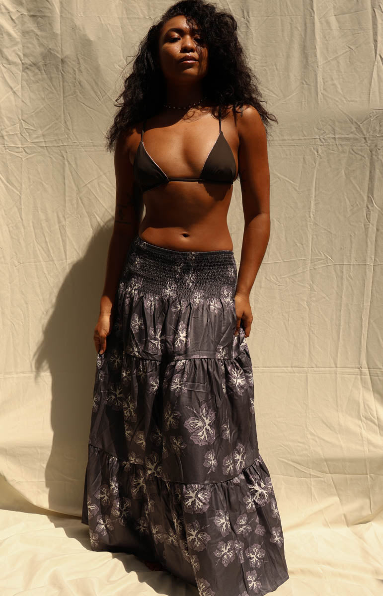 A woman modeling a dark black and gray tiered maxi skirt with a white floral pattern, paired with a black triangle bikini top, standing against a beige fabric backdrop. Tai Swim activewear skirt maxi dress