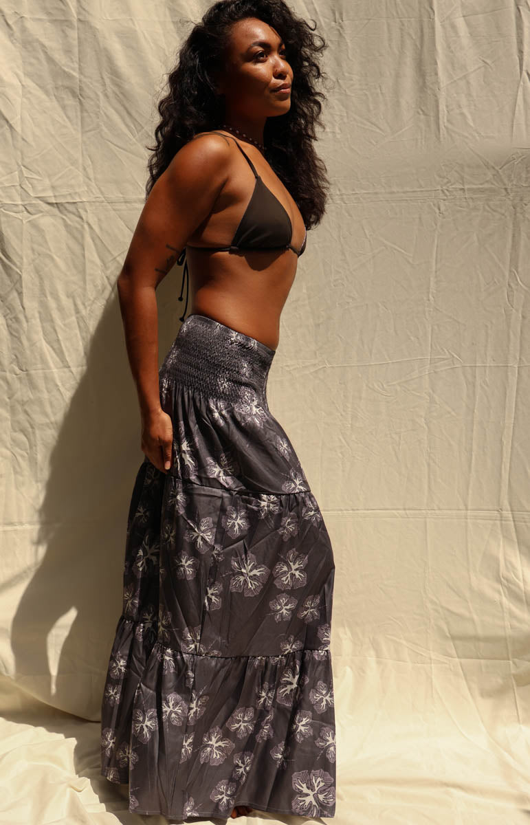 A woman modeling a dark black and gray tiered maxi skirt with a white floral pattern, paired with a black triangle bikini top, standing against a beige fabric backdrop. Tai Swim activewear skirt maxi dress