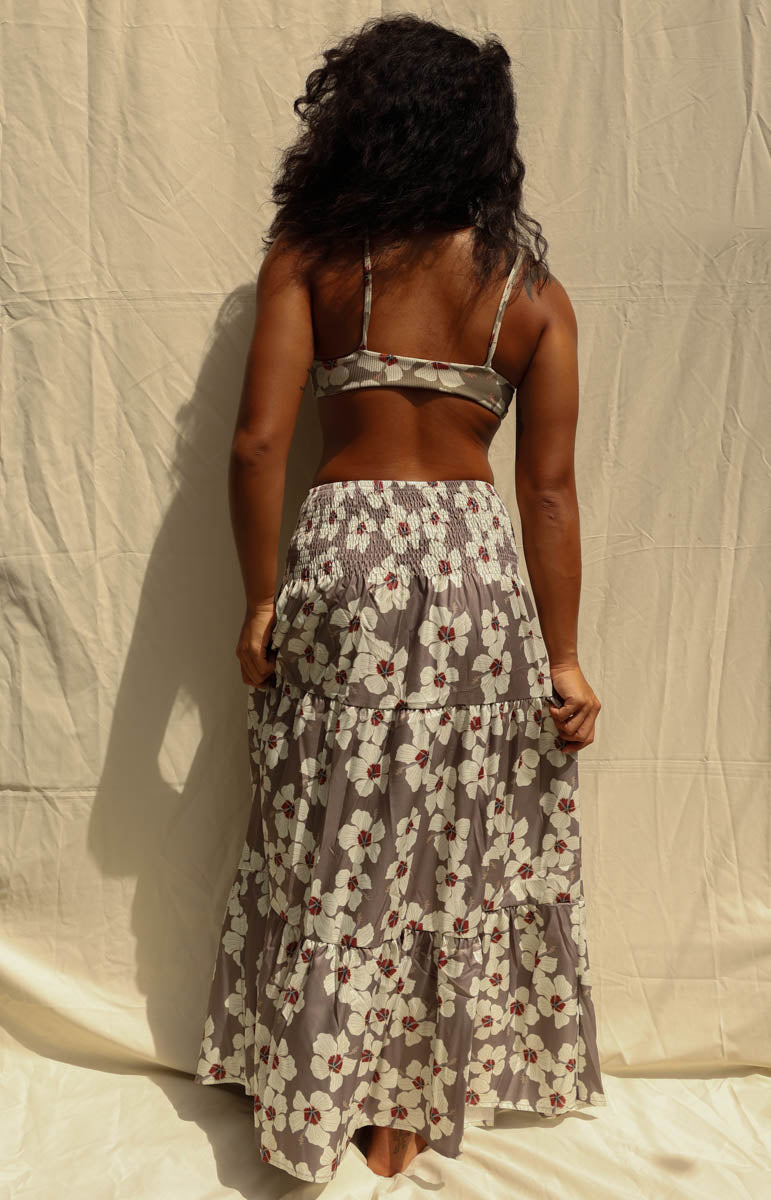 A woman modeling a matching two-piece floral set consisting of a wrap-style triangle crop top and a long tiered maxi skirt, in a gray, white, and red pattern, standing against a wrinkled beige fabric backdrop. Hibiscus floral from Kailua hawaiian brand tai swim